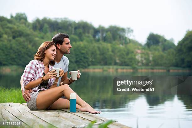 relaxed young couple with coffee mugs sitting on a jetty at lake looking at distance - steg zwei menschen stock-fotos und bilder