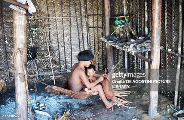 Yanomami woman with a baby on her lap building a straw basket inside a hut, Mato Grosso, Brazil.