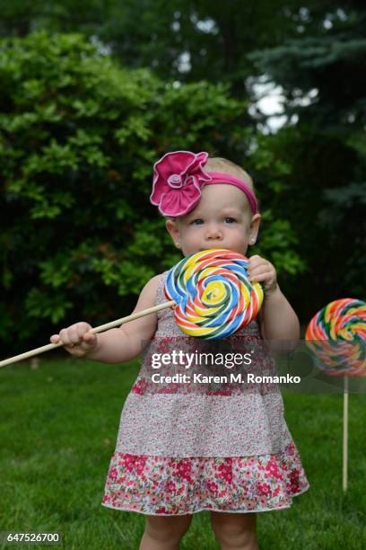 baby girl (14 mo.) standing outside; eating large swirl lollipop - eenjarig plantenkenmerk stockfoto's en -beelden