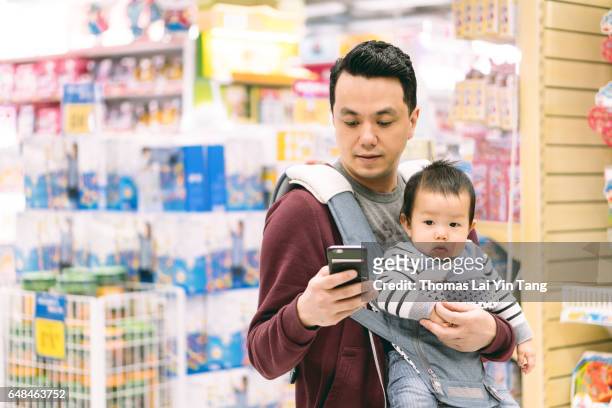chinese father checking mobile cell phone in supermarket while carrying 9 months old baby boy joyfully - baby carrier stock pictures, royalty-free photos & images