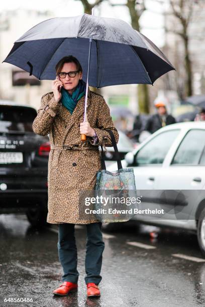 Hamish Bowles wears a scarf, a leopard print trenc coat, green pants, and red leather shoes, and an umbrella, outside the Celine show, during Paris...