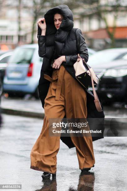 Guest wears a long black puffer jacket with a hood, a bag, and brown flared pants, outside the Celine show, during Paris Fashion Week Womenswear...