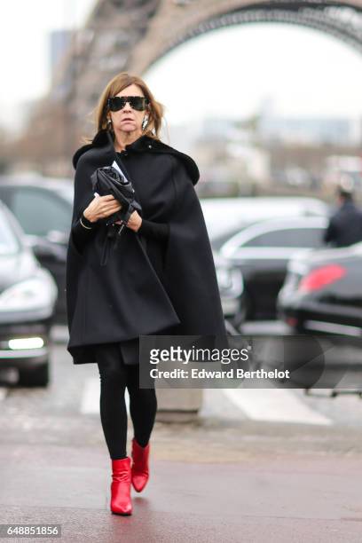 Carine Roitfeld wears sunglasses, a black cape, black leggings, and red leather boots, outside the Hermes show, during Paris Fashion Week Womenswear...