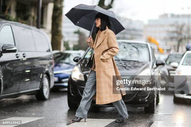 Guest wears gray pants, black heels, a trench coat, and an umbrella, outside the Celine show, during Paris Fashion Week Womenswear Fall/Winter...