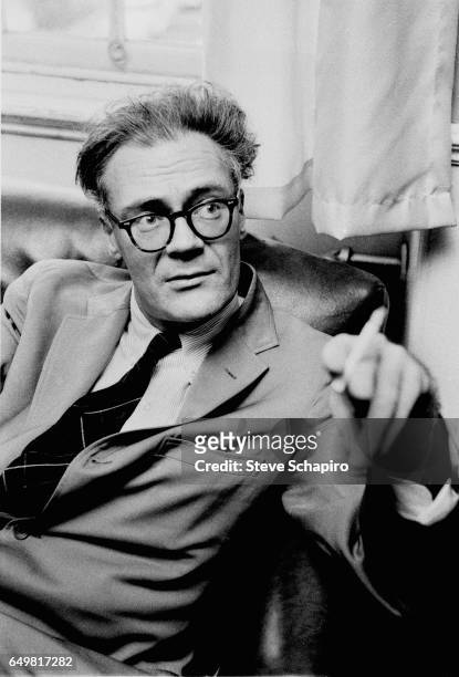 View of American poet Robert Lowell , a cigarette in his band, as he sits on a sofa, Boston, Massachusetts, 1964.