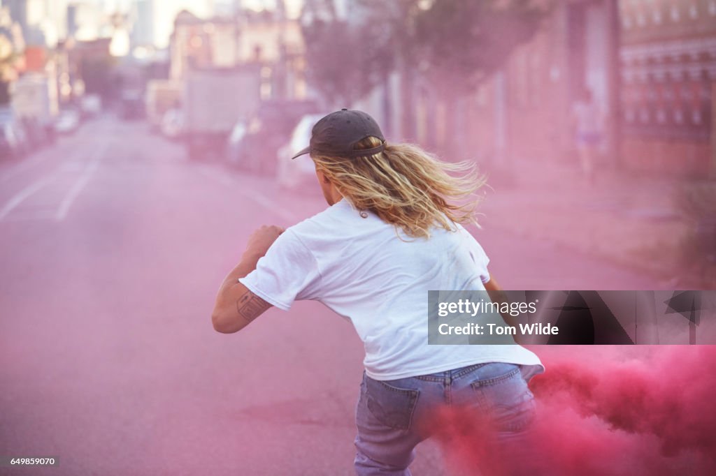 Young male skateboarder wearing white T-shirt carrying a pink flare as he skates away down the street