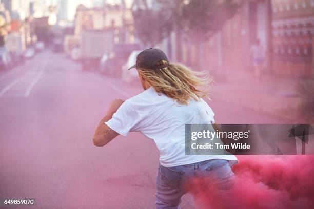 young male skateboarder wearing white t-shirt carrying a pink flare as he skates away down the street - insurrection stock-fotos und bilder