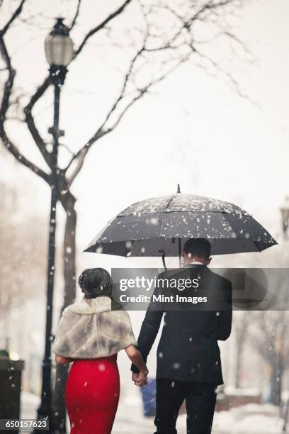 A woman in a long red evening dress and a man in a suit, walking through snow in the city.