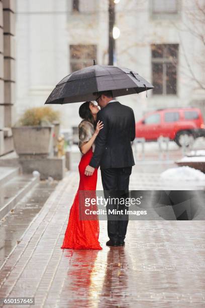 a woman in a long red evening dress with fishtail skirt and a fur stole, and a man in a suit kissing under an umbrella on a street. - wedding umbrella stock pictures, royalty-free photos & images