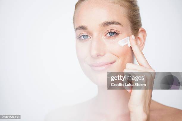 bare shouldered woman rubbing face cream into cheek looking at camera smiling - face cream stock pictures, royalty-free photos & images