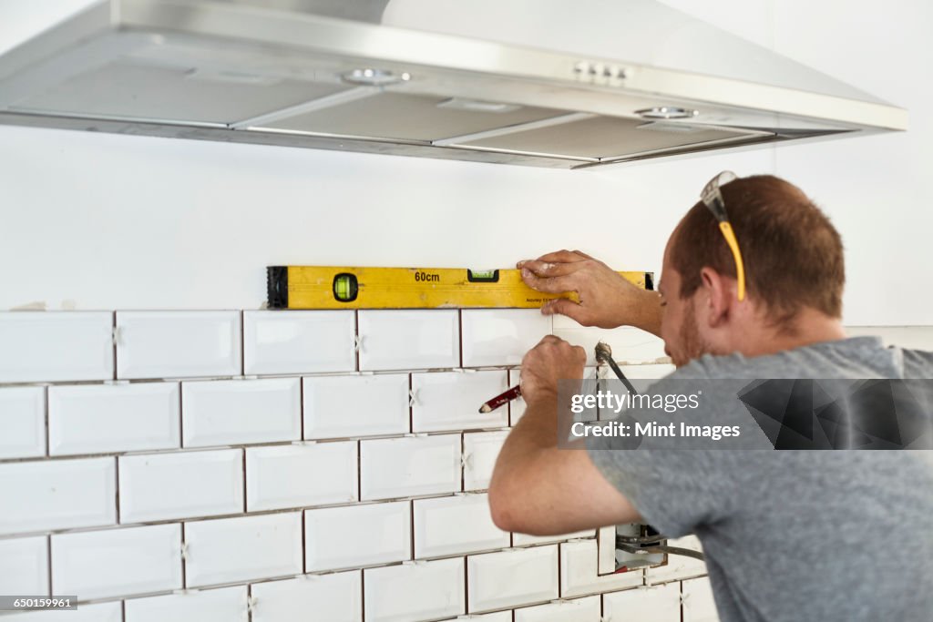A man working in a new kitchen, a tiler applying tiles to the wall behind the cooker.