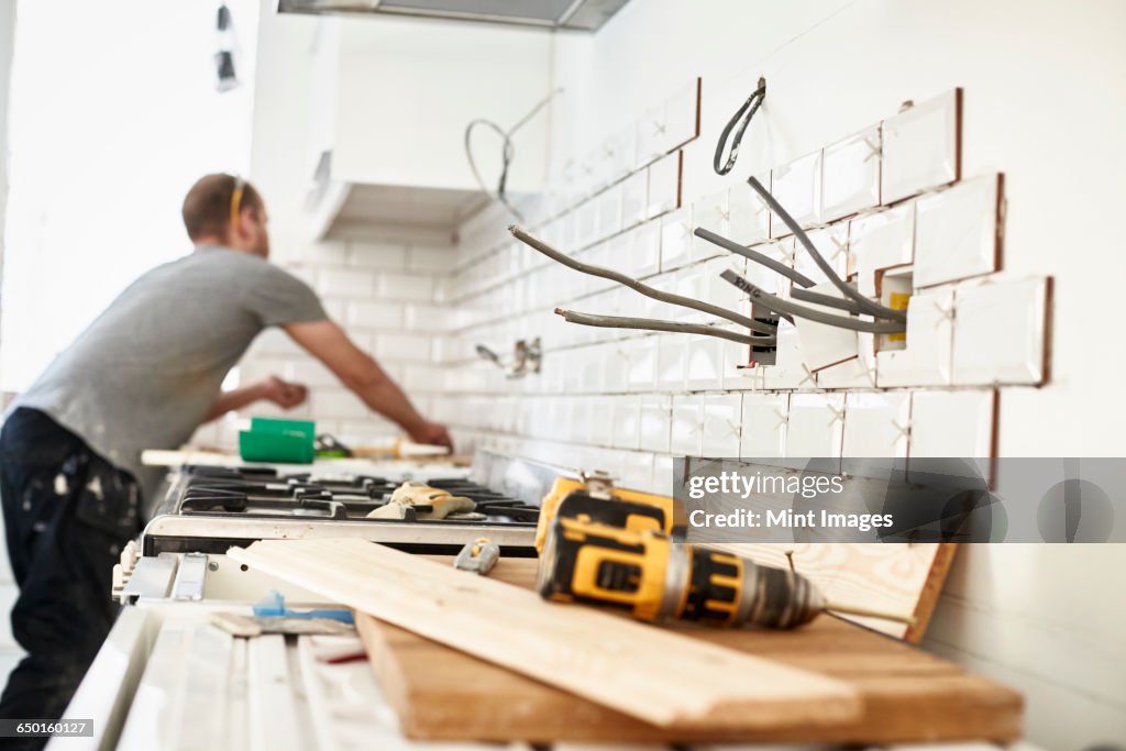 A man working in a new kitchen, a tiler applying tiles to the wall behind the cooker. 