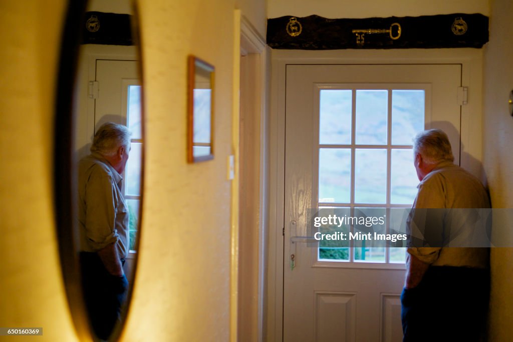 An elderly man standing by a door looking out through the window panes.