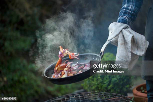 an outdoor cookout. a man holding a frying pan sauteeing vegetables above an open fire. - only mid adult men stock pictures, royalty-free photos & images