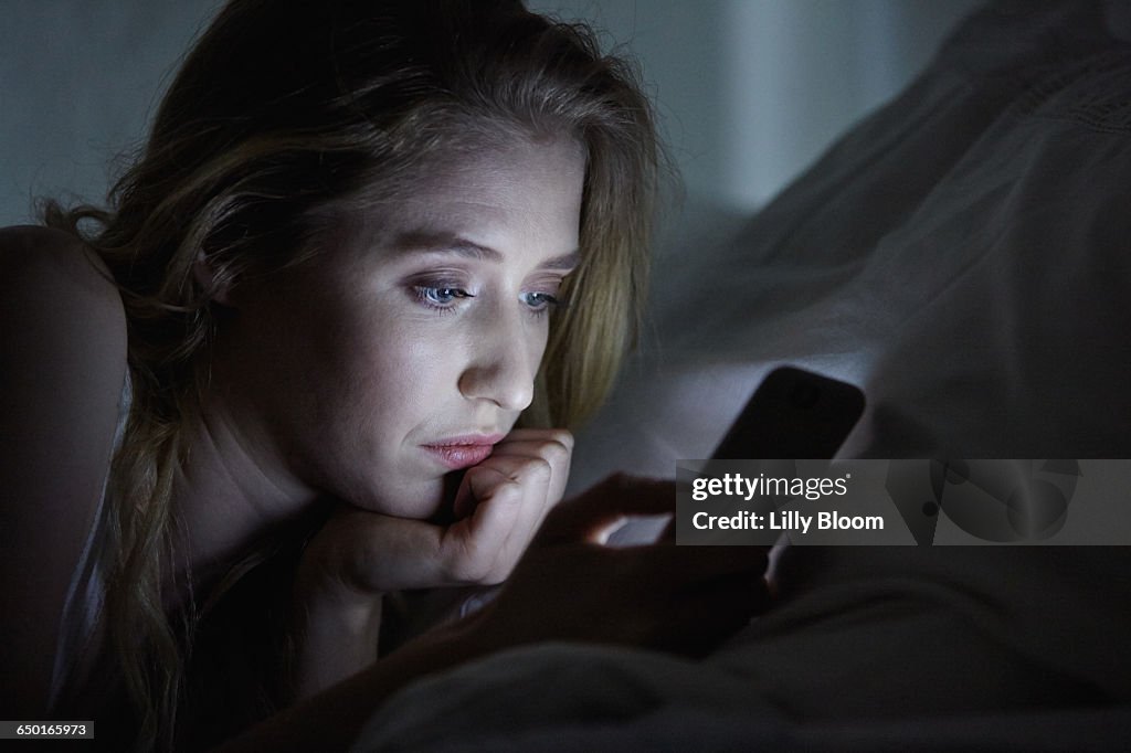 Close up of young woman lying in bed reading smartphone texts at night