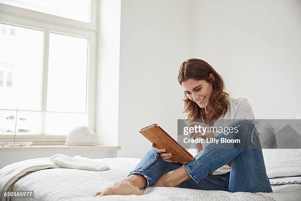 beautiful young woman sitting on bed using digital tablet - woman wearing white jeans stock-fotos und bilder