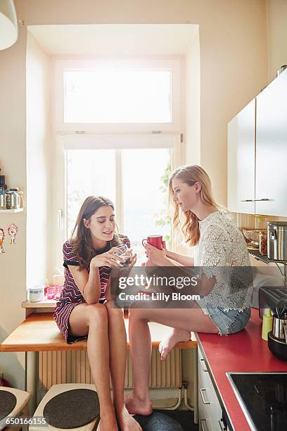 two young woman sitting on kitchen counter chatting - chica-tomando-cafe fotografías e imágenes de stock