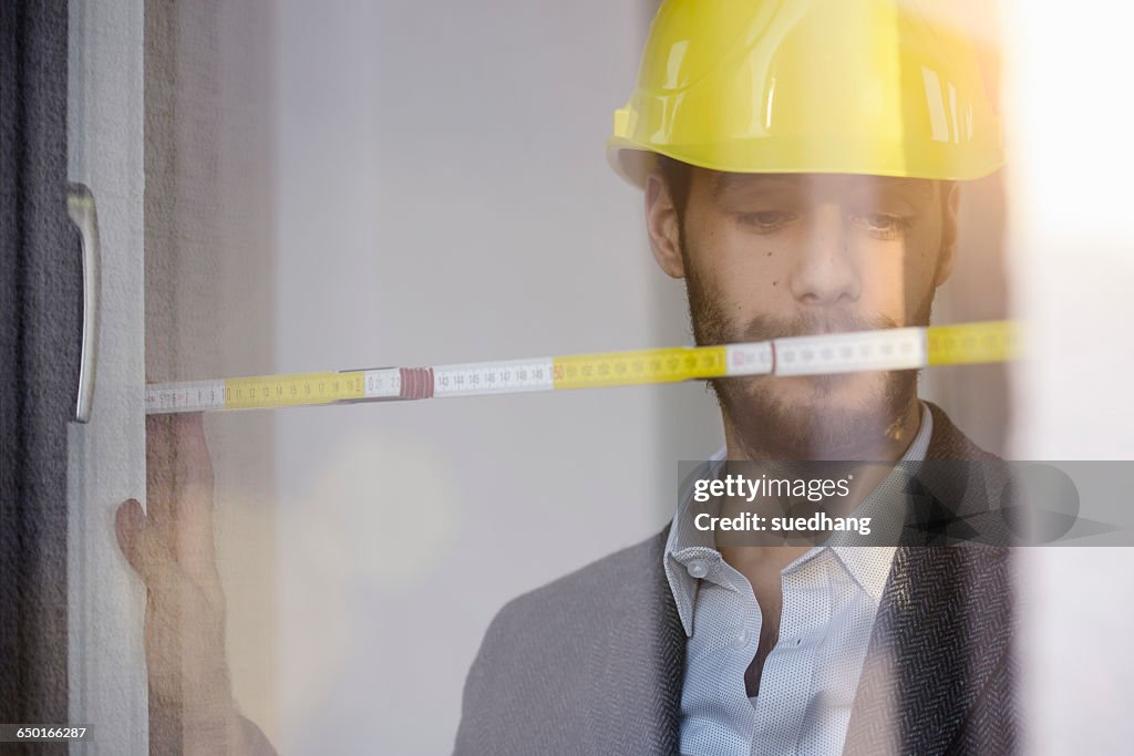 Young businessman wearing yellow hard hat measuring window in new office