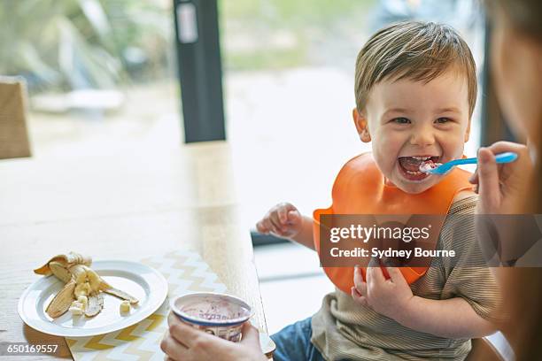 happy baby boy sitting at table being fed yogurt by mother - mother baby food stock pictures, royalty-free photos & images