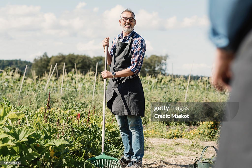 Mature farmer holding gardening fork and standing with coworker at farm