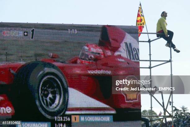 Ferrari fan, having climbed the TV scaffolding, sits with his flag whilst Michael Schumacher powers to pole position on the giant screen