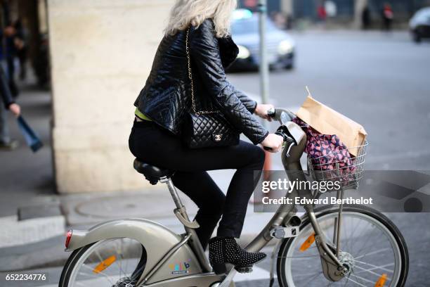 Passerby wears a black leather jacket, a Chanel bag, black pants, stud shoes, and is riding a bicycle, on March 11, 2017 in Paris, France.