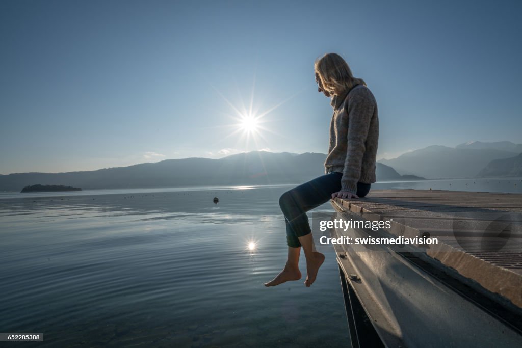 Young woman relaxes on lake pier, watches sunset