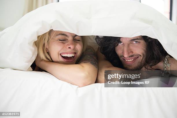 a cool young tattooed couple peeking out from under a duvet on a bed - couple under sheets ストックフォトと画像