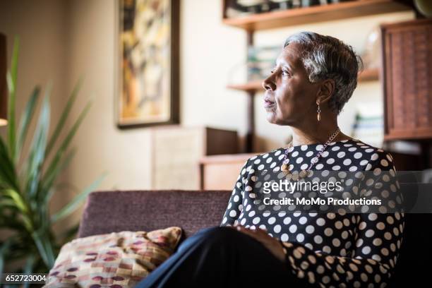 portrait of woman (60yrs) sitting on couch at home - woman concerned stockfoto's en -beelden