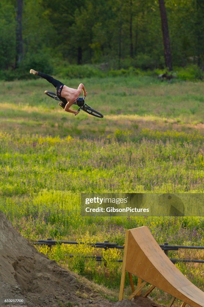 A young male mountain bike rider does a 360 nac-nac while riding off a wooden ramp jump.