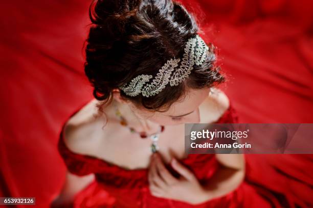 high angle studio shot of young woman in off-shoulder victorian red gown with dark hair in updo, looking down, surrounded by red satin - victorian gown fotografías e imágenes de stock