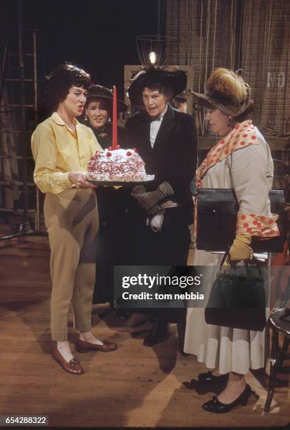 American actress Rosalind Russell holds a birthday cake with a red candle, as she stands with unidentified others backstage during the filming of the...