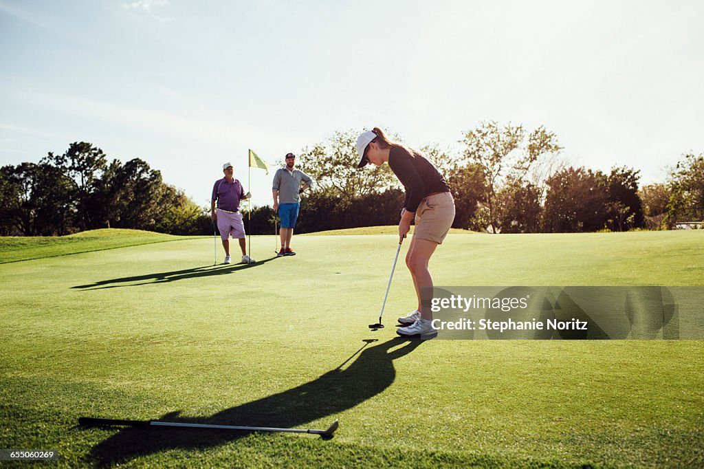 Woman Putting On Golf Course With Family Watching