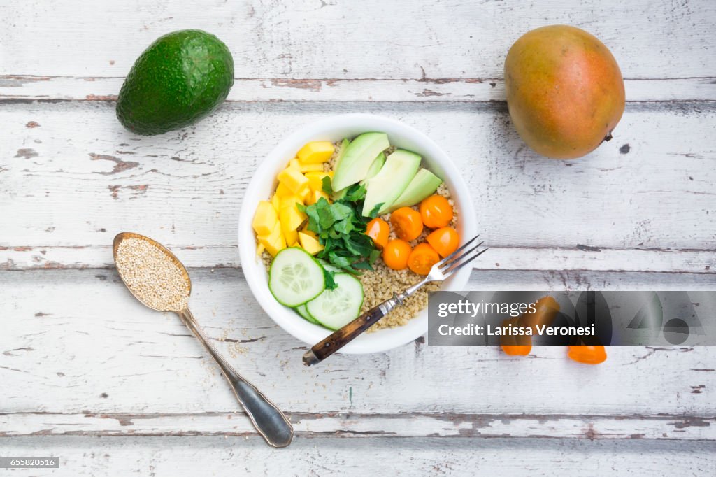 Lunch bowl of quinoa, mango, avocado, cucumber, orange tomatoes and parsley on wood