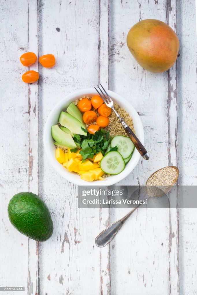 Lunch bowl of quinoa, mango, avocado, cucumber, orange tomatoes and parsley on wood