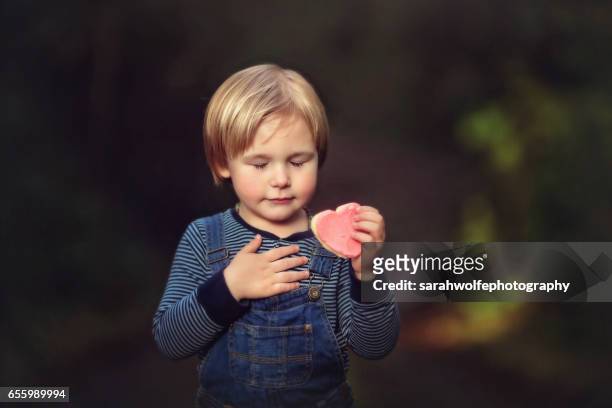 little boy touching heart while holding a heart shaped cookie - main sur la poitrine photos et images de collection