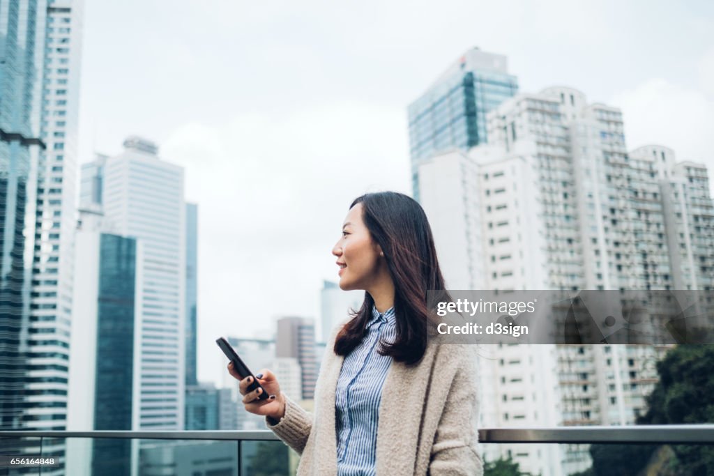 Young businesswoman reading emails on smartphone by the terrace of office building