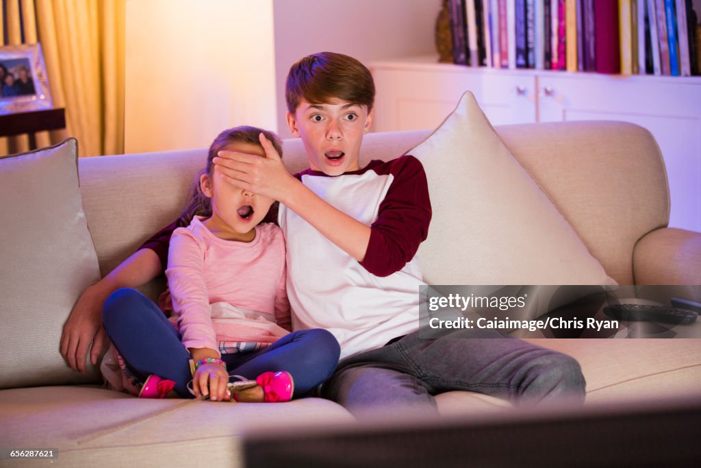 Brother covering surprised sisters eyes watching TV in living room