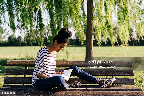 young woman sitting on park bench reading a book - mujer leyendo libro en el parque fotografías e imágenes de stock