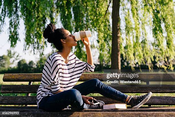 young woman sitting on park bench drinking coffee to go - banco del parque fotografías e imágenes de stock