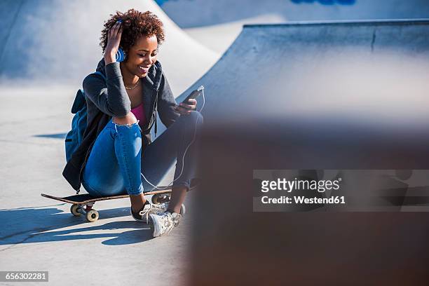 smiling young woman sitting on skateboard listening to music - skateboard park stock-fotos und bilder