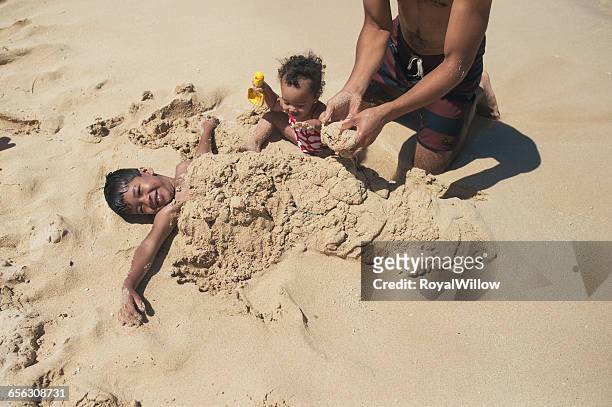 father and daughter burying son in sand on beach - buried in sand stock-fotos und bilder