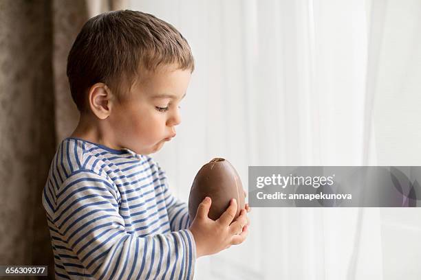 boy standing by window eating a large chocolate easter egg - eating chocolate easter egg stock pictures, royalty-free photos & images
