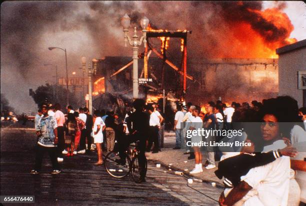 Rodney King Riot. Mother and child amidst mayhem, businesses burning, bystanders watching raging fires, pedestrian walking in the street drinking...