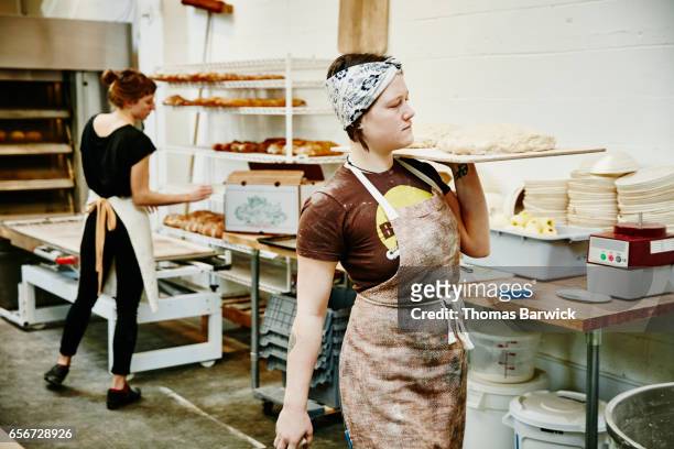 female baker carrying dough on pastry board through bakery - bäckerei stock-fotos und bilder