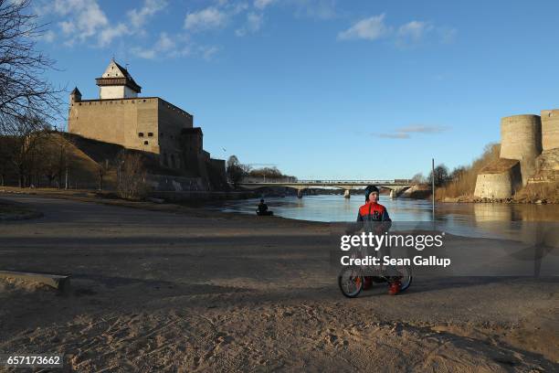 Russian-speaking boy pauses while riding his bike along a promenade along the Narva River below Hermann Castle and opposite Ivangorod Fortress, which...