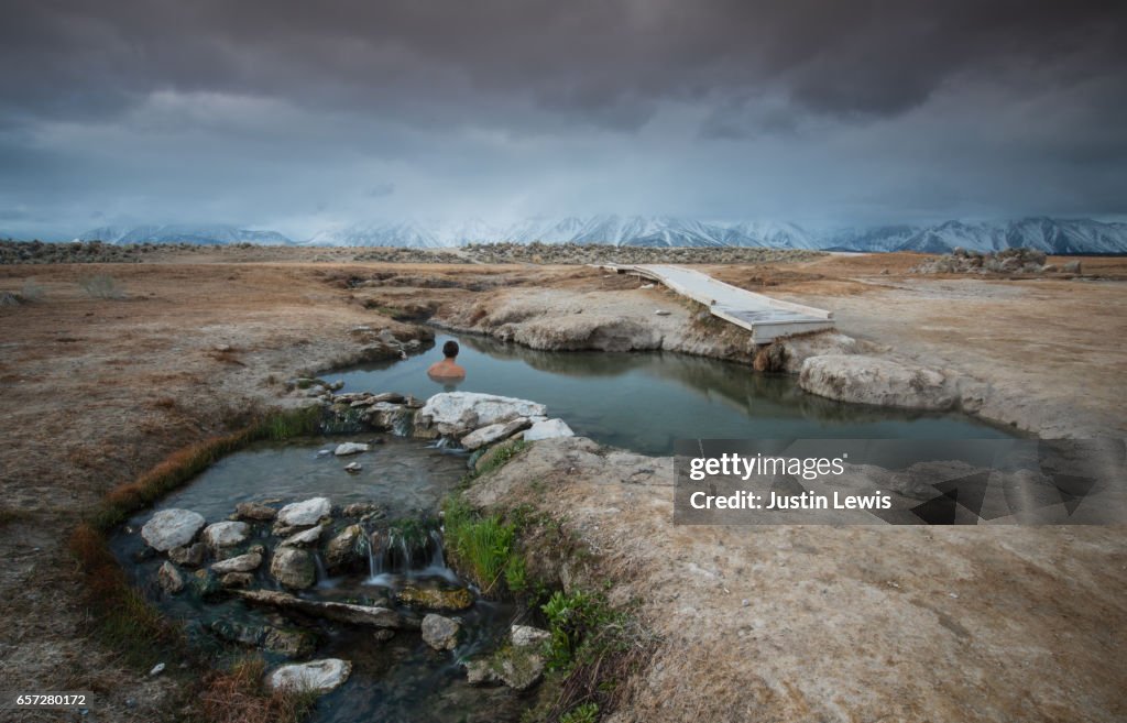 Solo Young Man Soaks in Natural Hot Springs in the Middle of a Vast Desert, Gazing at Horizon of Snow-capped Mountain Range, Flowing Water in Foreground
