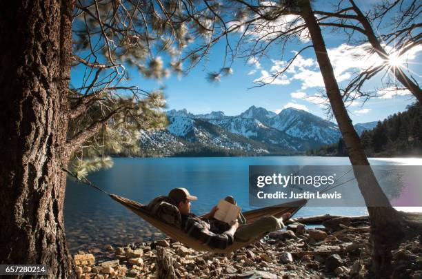 solo man backpacker reclines in hammock reading and relaxing by a mountain lake - travel with book stock pictures, royalty-free photos & images