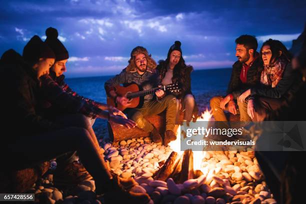 amigos tocando guitarra y cantando alrededor de la fogata en la playa - winter fire fotografías e imágenes de stock
