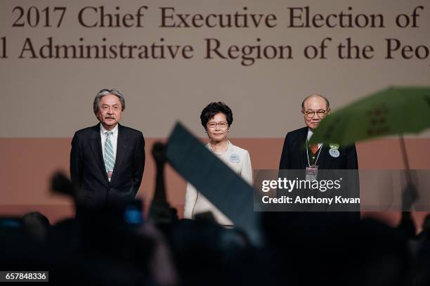Protesters hold a yellow umbrella and a banner as Carrie Lam, Hong Kong's chief executive-elect, center, stands on stage with candidates for Hong...